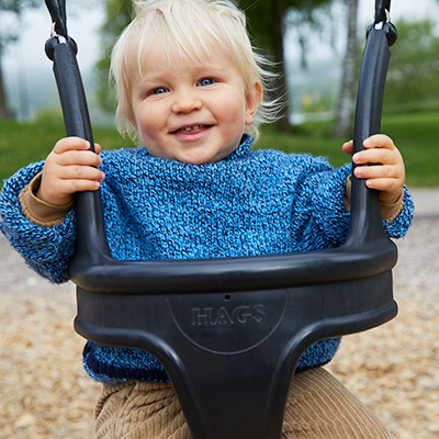 A young toddler child is swinging on a baby swing, looking directly at the camera and smiling.